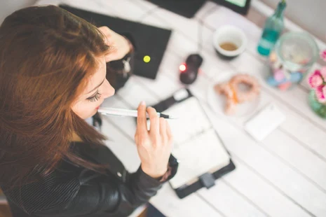 Eine Frau mit rotem Haar schreibt konzentriert in ein Notizbuch an einem Schreibtisch mit Laptop, Kaffee, Blumen und persönlichen Accessoires.