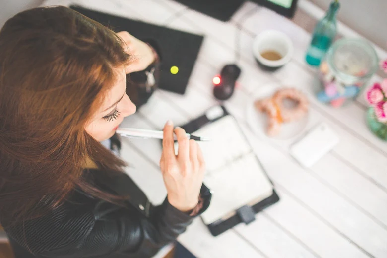 Eine Frau mit rotem Haar schreibt konzentriert in ein Notizbuch an einem Schreibtisch mit Laptop, Kaffee, Blumen und persönlichen Accessoires.