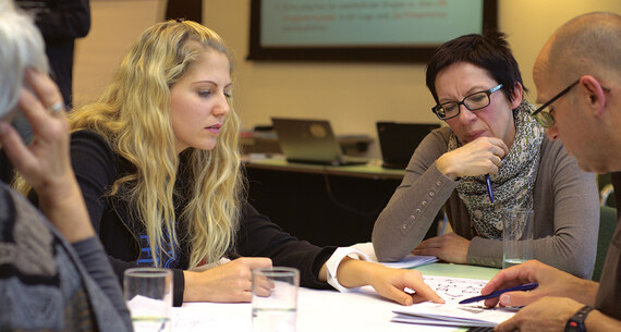 Personen sitzen an einem Tisch und lesen in Büchern. Eine blonde Frau erklärt den anderen etwas.
